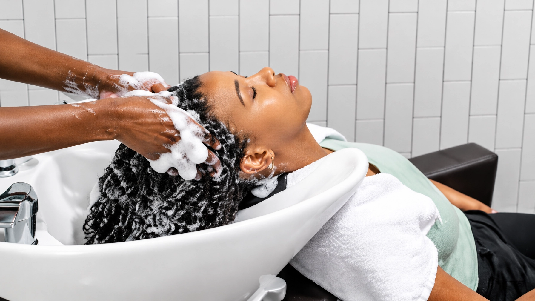A woman reclining at a salon wash basin with eyes closed while a stylist lathers her hair