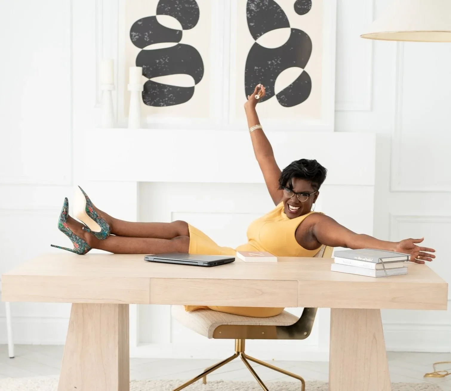 Natacha Pennycooke laughing in a yellow sleeveless dress and green-rimmed glasses while lying across a pale wood desk with her legs raised