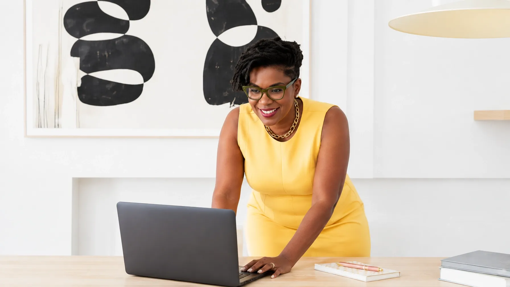 Natacha Pennycooke in a bright yellow sleeveless dress and green-rimmed glasses, leaning over a pale wood desk beside an open dark grey laptop