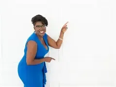 Natacha Pennycooke in a royal blue dress, smiling and pointing upward against a white wall