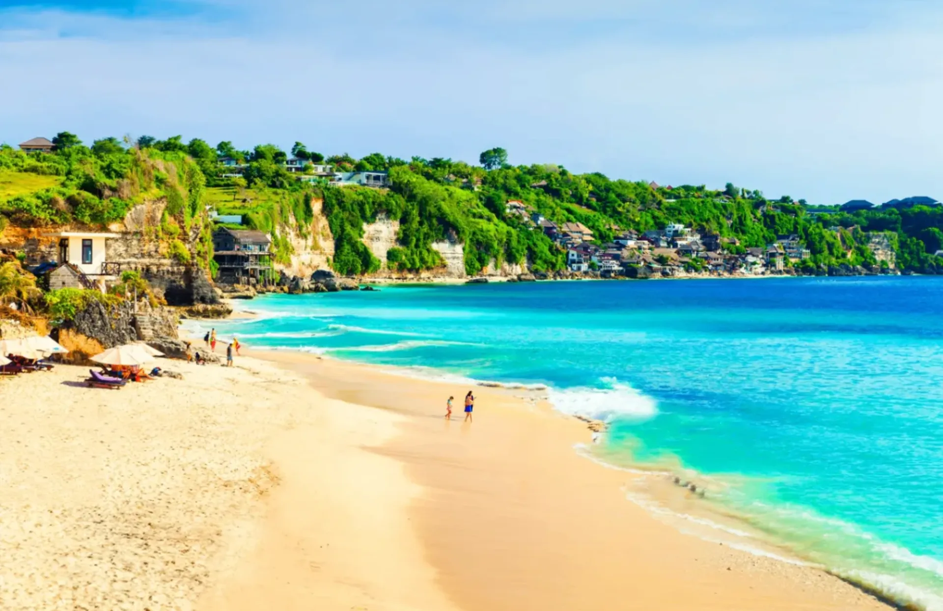 Wide Bali beach with golden sand, vivid turquoise water, green cliffs with villas behind, and blue sky