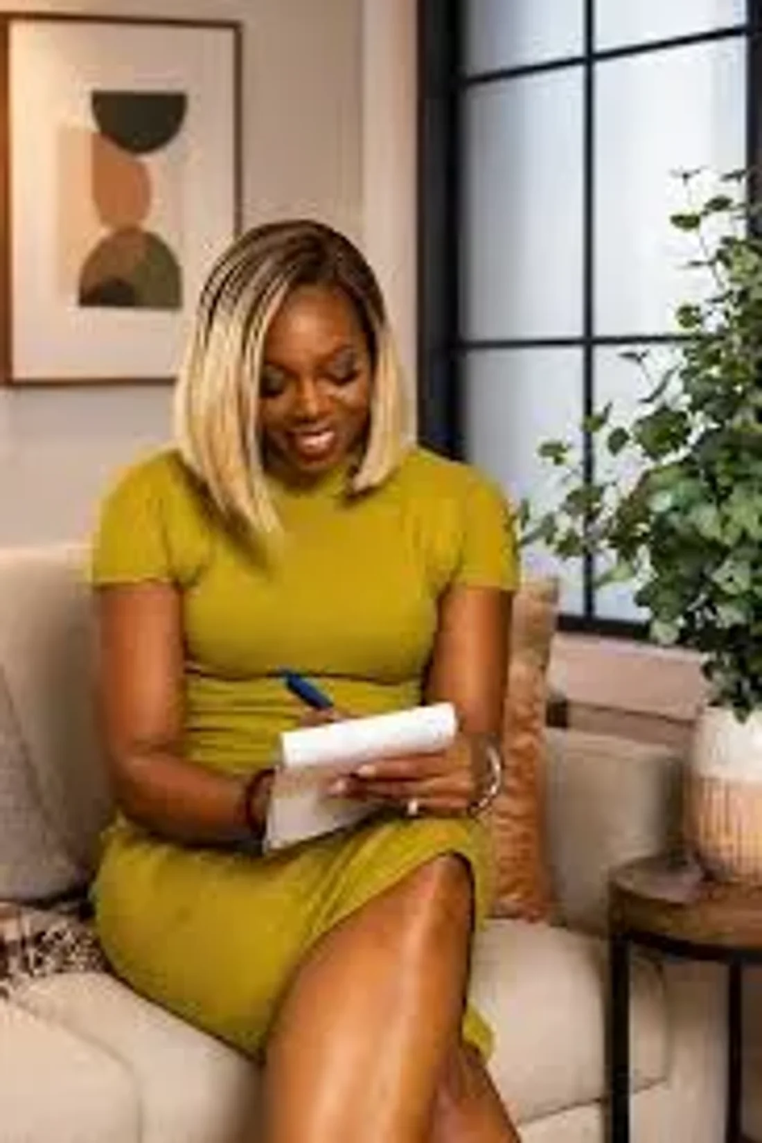 Eronmwon Balogun seated in a warm office in a chartreuse yellow dress, writing on a notepad with a blue pen