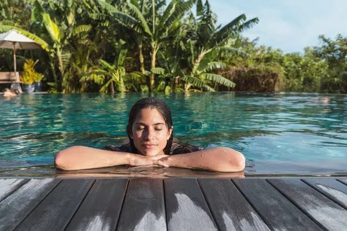 A woman resting her arms on the edge of a jungle infinity pool, eyes closed in calm relaxation surrounded by lush green palms
