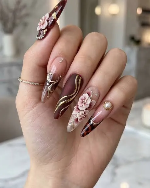 Burgundy stiletto nails with 3D pink floral art, gold swirls, pearls, and glossy nude accents