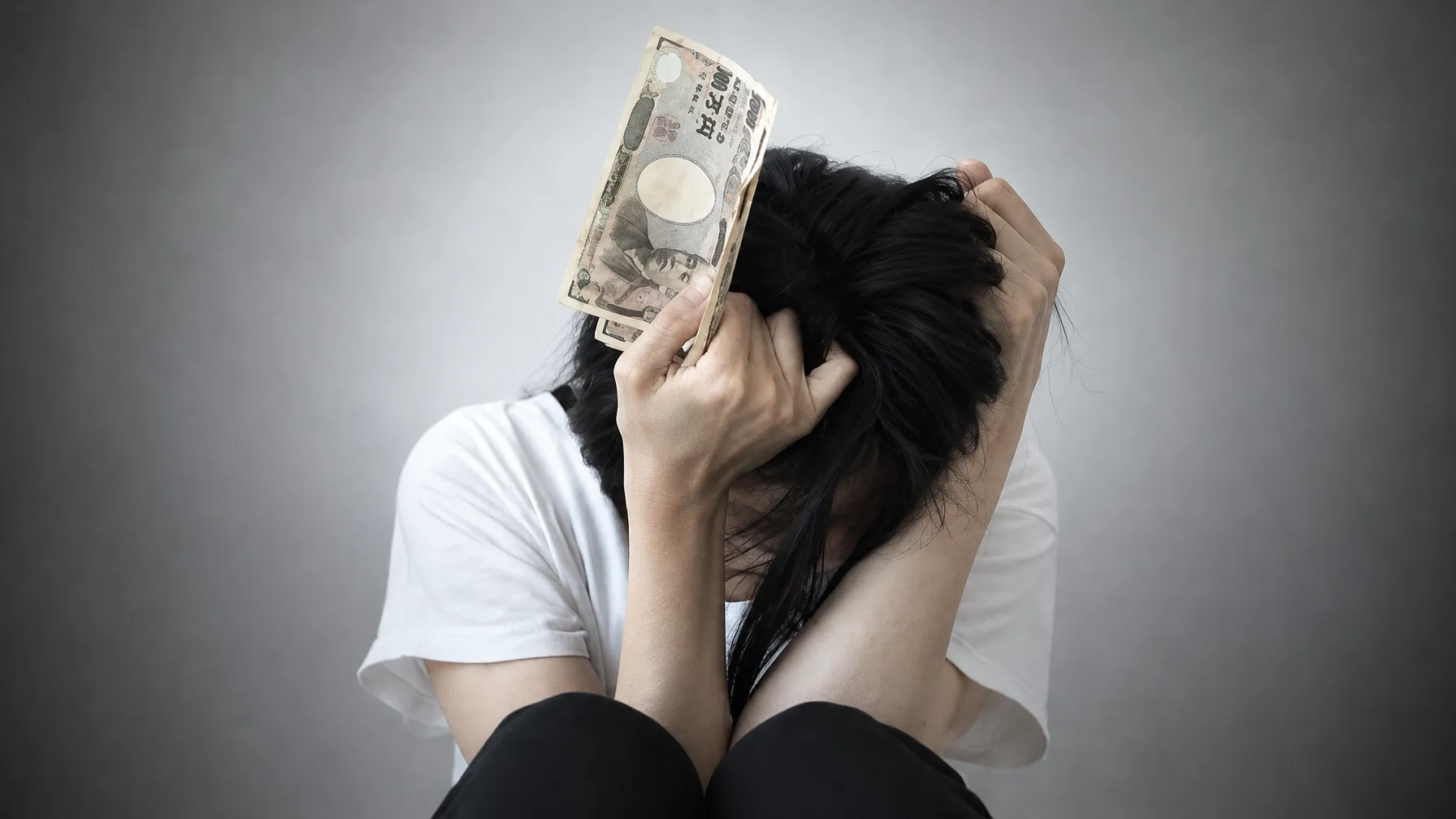 A woman sitting with her head bowed, one hand clutching her hair and the other holding a banknote